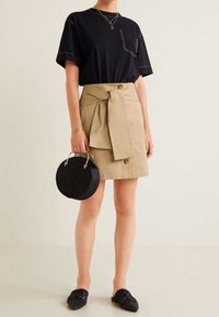 Camiseta negra de manga corta con bolsillo, falda beige con lazo y botones, bolso redondo negro de mimbre y zapatos oscuros tipo slip-on.