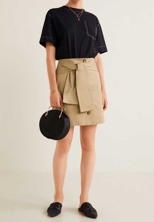 T-shirt noir à manches courtes avec poche, jupe beige avec ceinture à nouer et boutons, sac tissé noir rond, et chaussures noires à enfiler.