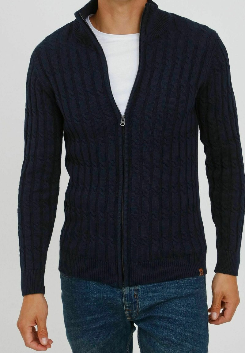 Mann in navyblauem, kabelgestricktem Zip-Pullover über einem weißen T-Shirt und blauen Jeans, steht mit entspannten Armen an den Seiten.