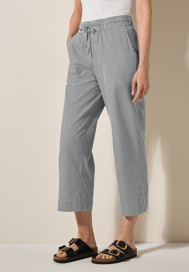 Personne portant un pantalon court rayé gris et blanc avec une taille à cordon et des sandales noires à double boucle, debout devant un fond beige.