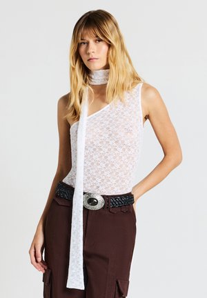 Donna che indossa un top bianco in pizzo a spalla unica con una sciarpa coordinata, pantaloni marroni a vita alta e una cintura nera intrecciata con fibbia argentata.