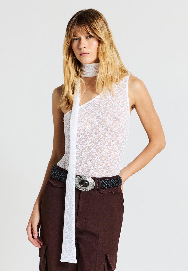 Donna che indossa un top bianco in pizzo a spalla unica con una sciarpa coordinata, pantaloni marroni a vita alta e una cintura nera intrecciata con fibbia argentata.