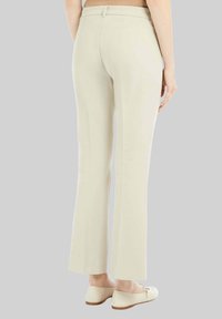 Donna che indossa pantaloni dritti color crema e scarpe flat abbinate color crema, in piedi contro uno sfondo grigio uniforme.