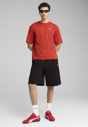 Rød bomulds t-shirt, sorte shorts, røde sneakers med hvide detaljer. Model med solbriller, står med armene bag ryggen foran en ensfarvet baggrund.