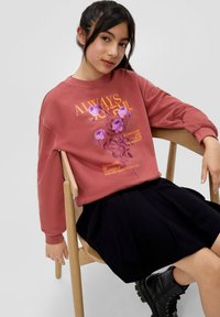 Bordeauxfarbener Pullover mit floralem Grafikdesign und Text, kombiniert mit einem schwarzen, plissierten Rock und schwarzen Kampfstiefeln, sitzt auf einem Holzstuhl.