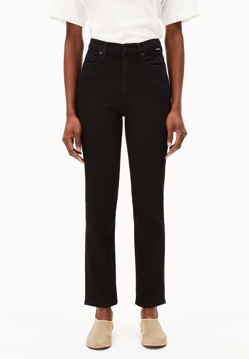 Personne portant un jean noir taille haute à jambes droites, un haut blanc à manches courtes et des chaussures beiges faciles à enfiler, debout devant un fond blanc.