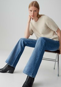 Lichtbeige, kortalige, harige trui gecombineerd met blauwe flared jeans en zwarte hakken enkellaarsjes. Model zittend op een metalen stoel.