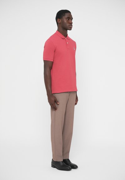 Punane lühikeste varrukatega polosärk, millel on väike logo. Seljas beežid püksid ja mustad kingad, seistes valge tausta ees.