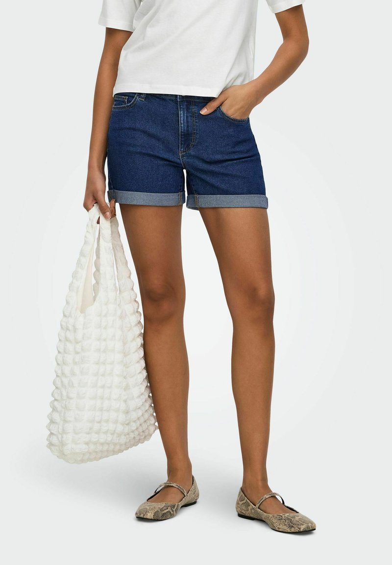 Femme portant un short en jean foncé roulé, un t-shirt blanc court, des ballerines imprimé serpent, et tenant un sac tote blanc texturé.