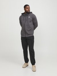 Grijze zip-up hoodie met een trekkoordcapuchon en een borstzak, gecombineerd met zwarte cargobroek en lichtbeige schoenen. Glad stoftextuur.