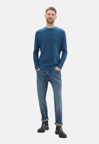 Suéter azul de punto con cuello redondo, combinado con jeans azul claro. El modelo lleva botas negras hasta el tobillo; manos en los bolsillos. Fondo minimalista.