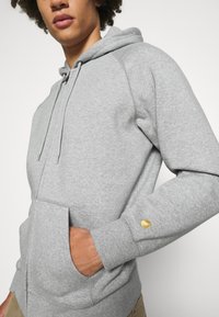 Grauer Zip-Hoodie aus Baumwollmischung, mit einer Kängurutasche und gerippten Bündchen, sowie einem kleinen goldenen Logo nahe dem linken Handgelenk.