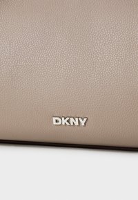 Beige skinnveske med en teksturert overflate, med en sølvfarget DKNY-logo i fet skrift. Nærbilde fremhever materialdetaljer.