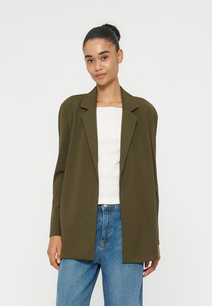 Femme aux cheveux foncés en chignon portant un blazer vert olive, un haut blanc et un jean bleu, debout devant un fond clair uni.