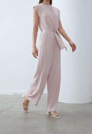Femme marchant dans une combinaison sans manches rose pâle avec jambes larges et ceinture à la taille, portant des sandales blanches à talons blocs à lanières sur un sol gris.