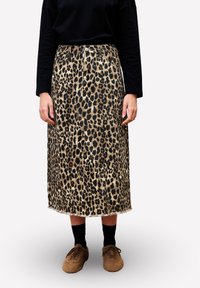 Personne portant une jupe midi imprimée léopard, un haut noir à manches longues, des chaussettes noires et des chaussures à lacets marron se tenant contre un fond blanc.