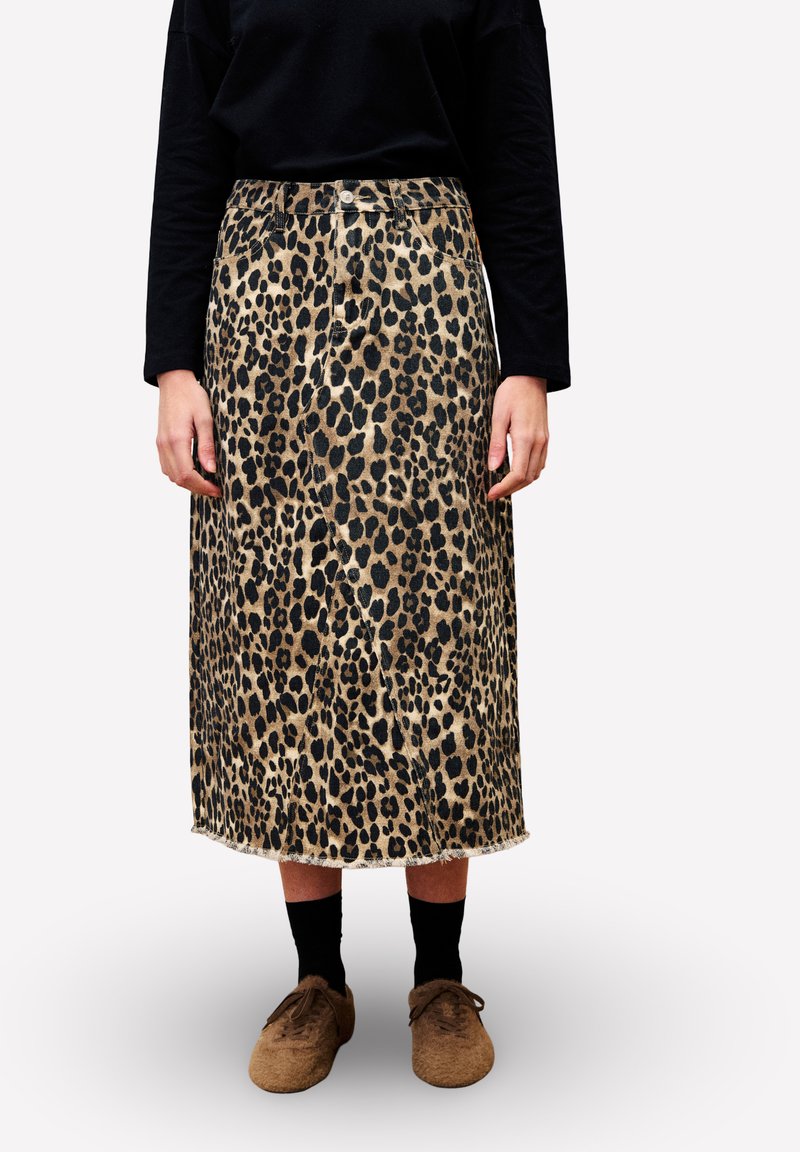 Personne portant une jupe midi imprimée léopard, un haut noir à manches longues, des chaussettes noires et des chaussures à lacets marron se tenant contre un fond blanc.