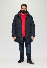 Marineblaue Steppjacke mit gesteppter Oberfläche und Kapuze, ausgestattet mit rotem Innenfutter und zwei Reißverschluss-Brusttaschen. Kombiniert mit einem roten Shirt und Jeans.