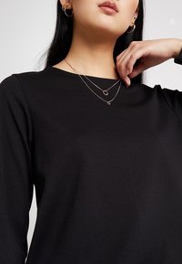 Černý top s dlouhými rukávy a kulatým výstřihem. Modelka nosí dvě zlaté vrstvené náhrdelníky a malé kruhové náušnice. Hladká textilie.