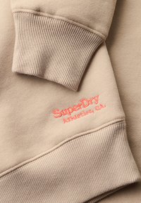 Beige sweatshirt med ribbede manchetter. Har koralfarvet broderet tekst "SuperDry Athletics, C.A." på forsiden. Blødt, tekstureret stof.