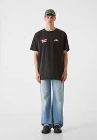 Prevelik temno siv bombažni T-majica z rožnato besedilom, v kombinaciji s svetlo modrimi širokimi hlačami in črnimi platformskimi čevlji. Nosena je preprosta zelena kapa.