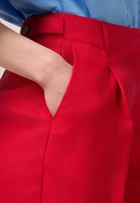 Rode, geplooide broek met een gestructureerde afwerking, voorzien van een zijzak. Gedetailleerd met een gladde tailleband en een knopenaccent.