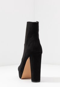 Czarne zamszowe botki za kostkę z wysokim słupkowym obcasem, zaokrąglonym noskiem oraz bezszwowymi wykończeniami. Miękka tekstura z beżową podeszwą i minimalnym zdobieniem.