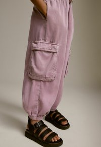 Ljust lila bomullscargobyxor med elastiska ankle cuffs, sidofickor och detaljerade sömmar, tillsammans med svarta sandaler med remmar.