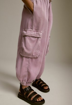 Ljust lila bomullscargobyxor med elastiska ankle cuffs, sidofickor och detaljerade sömmar, tillsammans med svarta sandaler med remmar.
