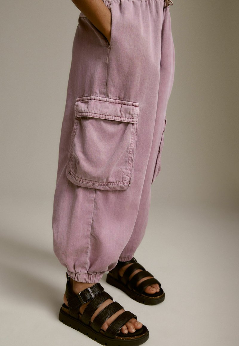 Ljust lila bomullscargobyxor med elastiska ankle cuffs, sidofickor och detaljerade sömmar, tillsammans med svarta sandaler med remmar.