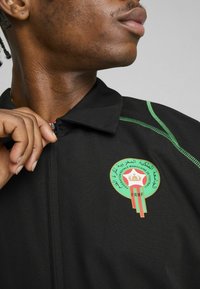 Sort jakke med en grøn lynlås og syninger, der har et rundt marokkansk fodboldforbunds badge på brystet.