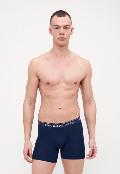 Námornícke modré boxerky s kontrastným logom na páse, priliehavý strih a hladká textúra tkaniny, modelované bez trička na mužskej postave.