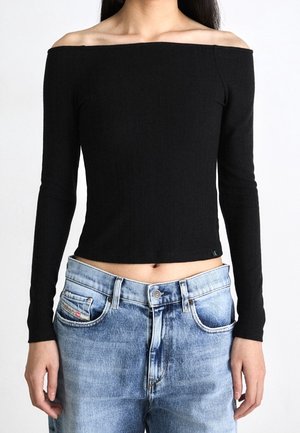 Vrouw draagt een nauwsluitende zwarte off-shoulder top met lange mouwen en lichtblauwe hooggehuisde jeans, de handen ontspannen langs de zijkant.