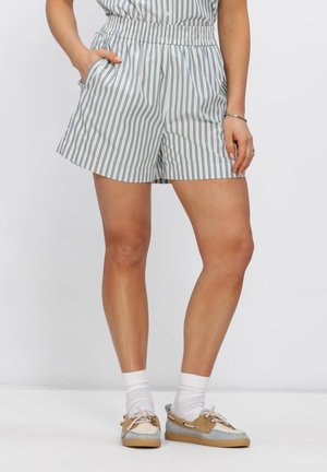 Vrouw die witte en blauwe shorts met verticale strepen draagt, witte sokken en beige en blauwe bootschoenen, staand met één hand in haar zak.