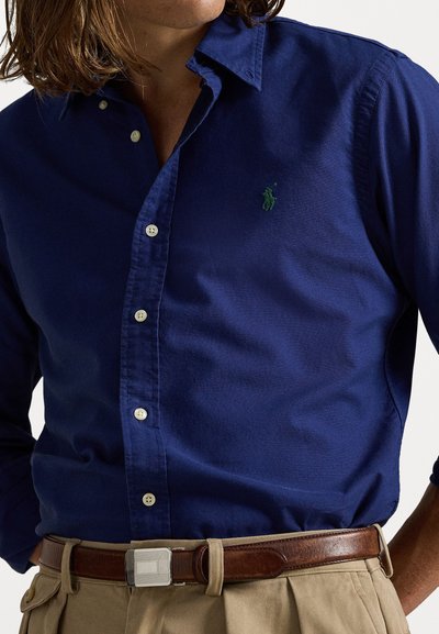 Navy zils krekls ar pogām, kas izgatavots no kokvilnas, ar mazu zaļu iz�šūtu logotipu, gaišām pogām un brīvu apkakli.