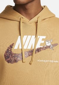 Hnědá mikina s kapucí s velkým bílým logem Nike a designem kávového zrnka. Pod tím je text "Součást vaší každodenní rutiny." S nastavitelnými šňůrami.