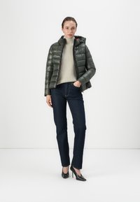 Veste verte rembourrée avec capuche, en tissu texturé et fermeture éclair, portée sur un pull gris clair et un jean noir, complétée par des talons noirs.