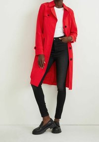 Abrigo trench rojo con botones negros, llevado sobre una camiseta blanca y jeans skinny negros. Los zapatos son negros con una suela gruesa.