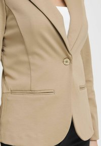 Blazer beige con chiusura a un bottone, rever inclinati e due tasche frontali; realizzato in tessuto testurizzato. Design aderente.