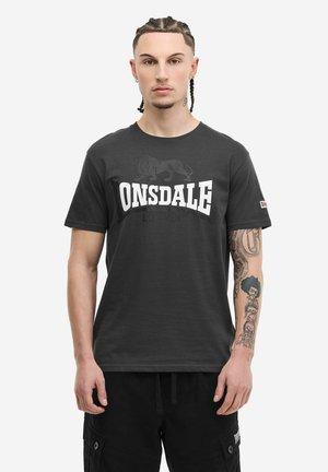 Man with braided hair and tattoos wearing a black Lonsdale T-shirt and black cargo shorts against a plain white background.