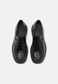 Mocassins noirs avec une finition brillante, présentant une bande à l'avant et des semelles en caoutchouc texturé. Tige en cuir lisse, design minimaliste.