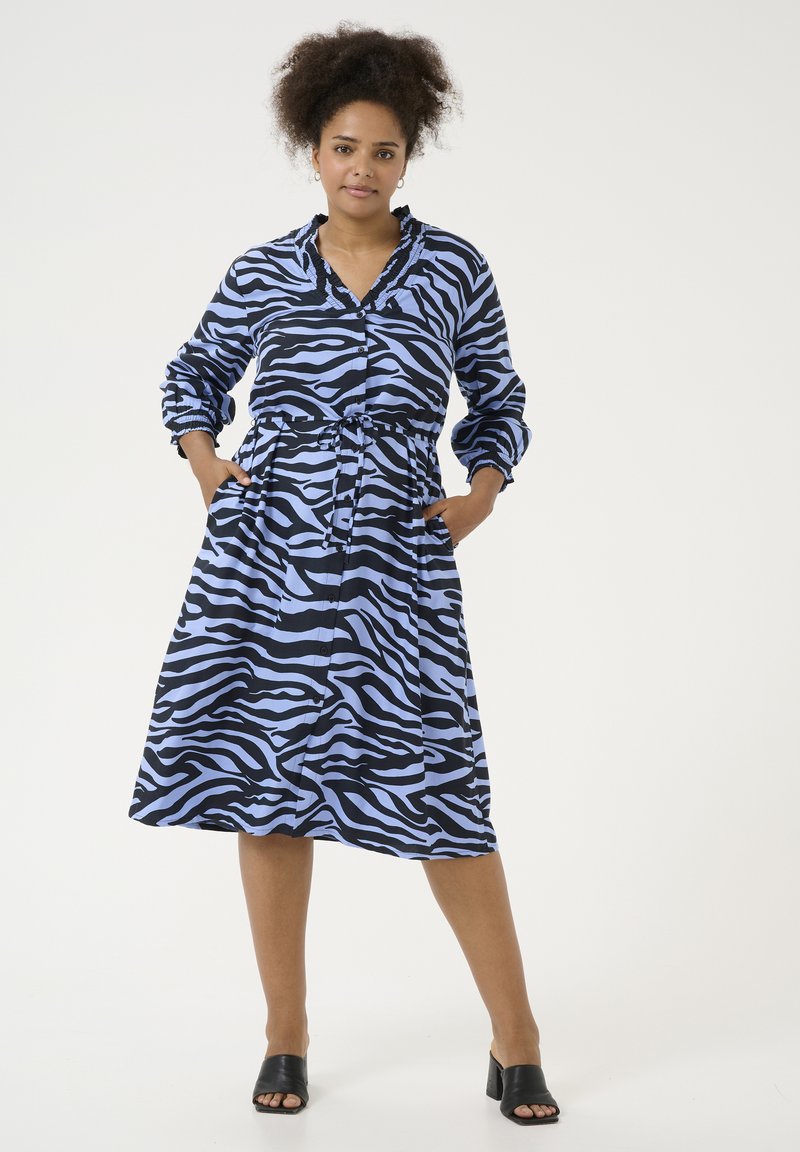 Zebra-print jurk in zwart en lichtblauw, met knoopsluiting aan de voorkant, lange mouwen, getailleerde taille, knielang. gedragen met zwarte schuifsandalen.