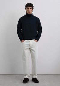 Jersey de cuello alto azul oscuro, pantalones rectos blancos y zapatos de ante marrón; modelo de pie con las manos en los bolsillos frente a un fondo neutro.