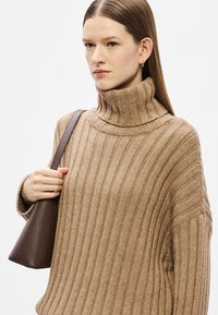 Μπεζ ριμπ πουλόβερ με ψηλό γιακά, απαλή υφή και μακριά μανίκια. Στο αριστερό χέρι κρατείται μια σκούρα καφέ τσάντα ώμου.