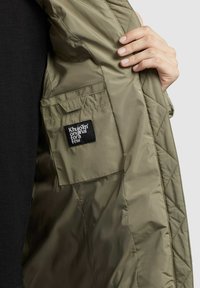 Interior de chaqueta acolchada color verde oliva con un bolsillo izquierdo; presenta una etiqueta negra con texto blanco. Textura de tela sintética suave.