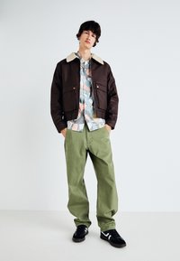 Braune Bomberjacke mit einem cremefarbenen Kunstpelzkragen, gemustertes Hemd in Pastellfarben, grüne weit geschnittene Hose und schwarze Schuhe mit weißen Akzenten.
