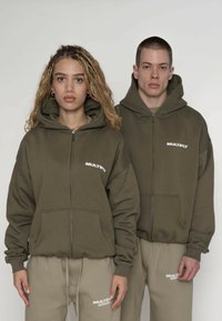 Sweat-shirts à capuche zippés vert olive avec poches avant, arborant un logo blanc "MULTIPLY". Associés à des joggeurs beige avec des accents de logo.