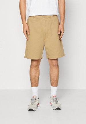 Shorts en khaki avec une coupe décontractée, fermeture à bouton et zip, deux poches latérales et une texture en coton lisse. Accompagnés de chaussures de sport grises.