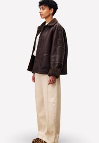Femme se tenant de côté, portant une veste en shearling marron foncé, un pantalon large crème et des chaussures à motif léopard contre un fond uni.
