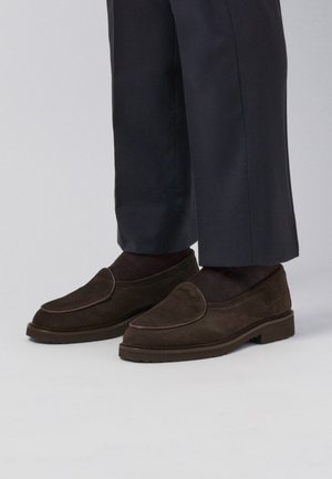Braune Wildleder-Slipper mit runder Zehenform und dekorativen Nähten. Dicke Gummisohle mit strukturiertem Griff. Kombiniert mit dunklen Hosen und Socken.